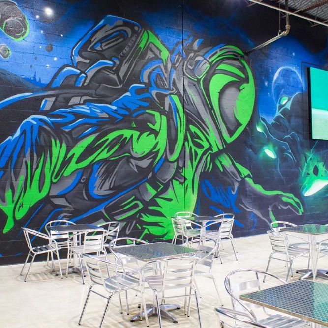Green and blue graffiti astronaut painted along a cinder block wall above cafe seating