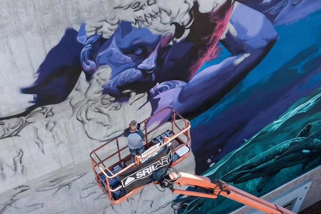 Aerial view of a monumental blue figure mural, the artist painting it from a boom lift