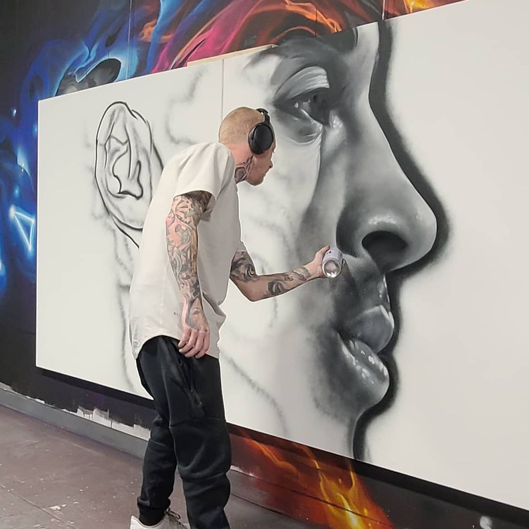 Hand-drawn underdrawing still visible beside the finished half of a large grayscale face mural, artist mid-spray