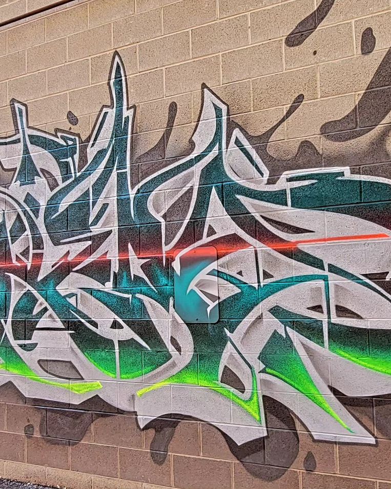Wildstyle graffiti lettering in silver, teal and green spray paint across a tan brick wall