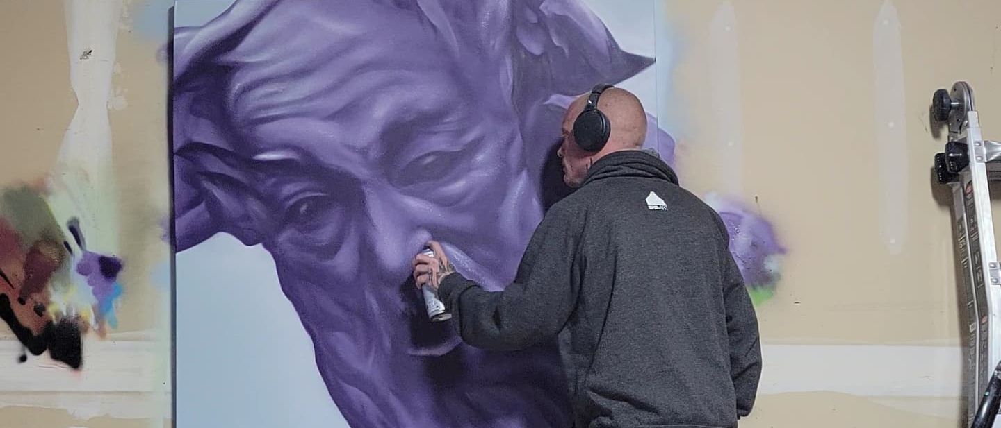 Spray-painting a large purple portrait onto a canvas propped against a studio wall