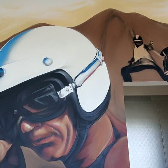 Hand-painted portrait mural of a helmeted motorcycle racer beside a stunt-rider scene on an interior wall