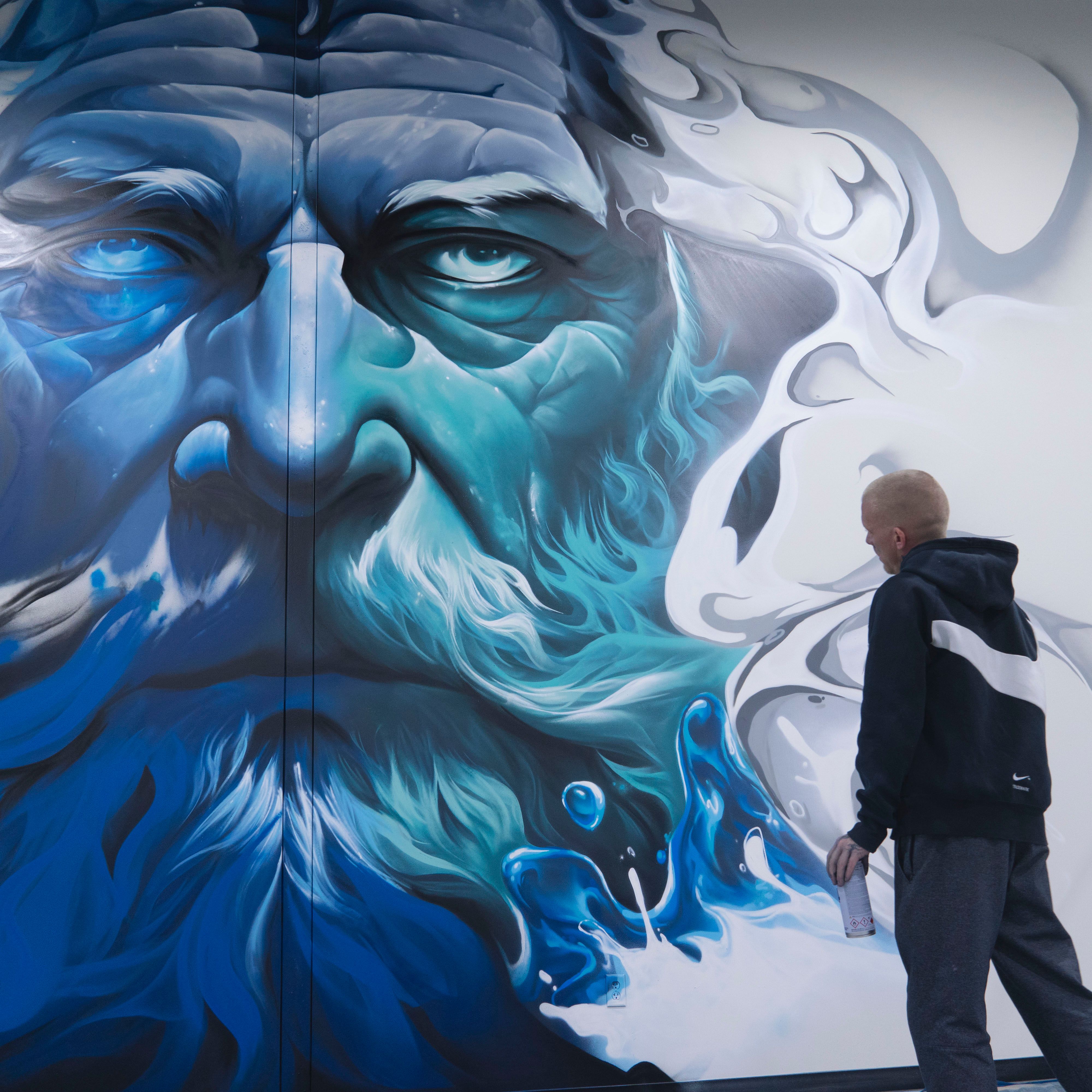 Monumental Zeus face mural in blue-toned grayscale, the artist standing before the nearly finished wall