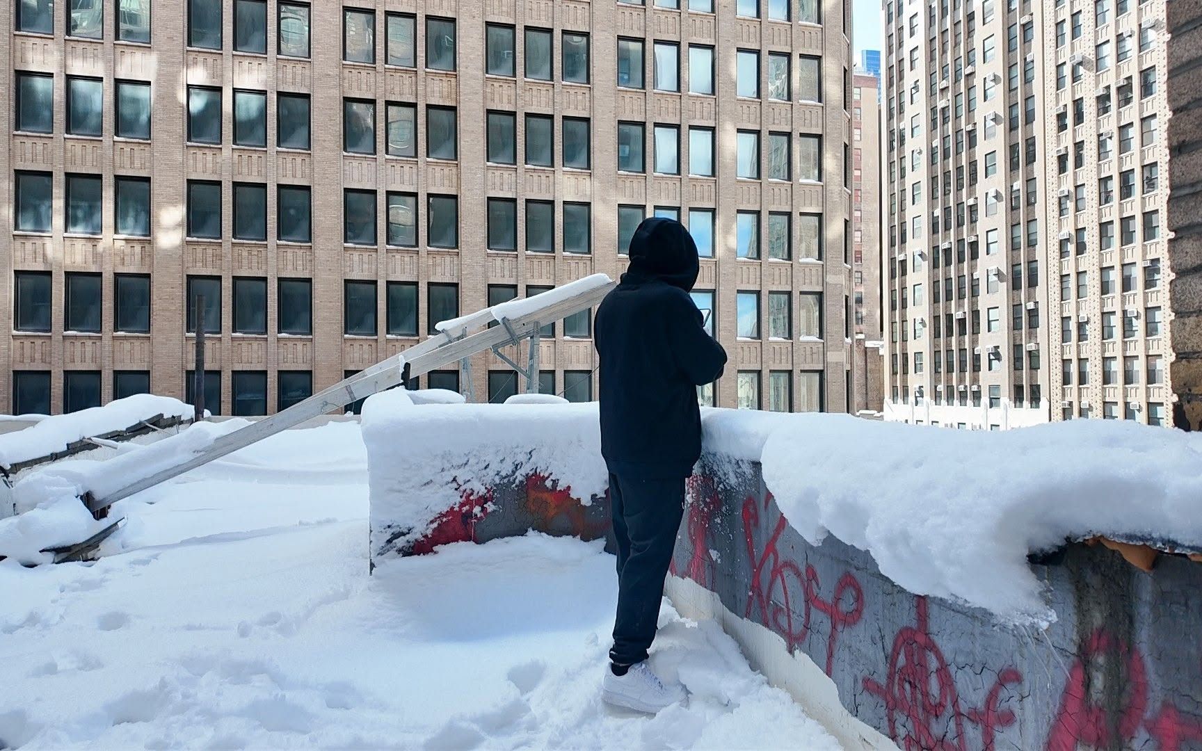 Standing on a snow-covered Manhattan rooftop beside a ladder, brick towers rising on every side