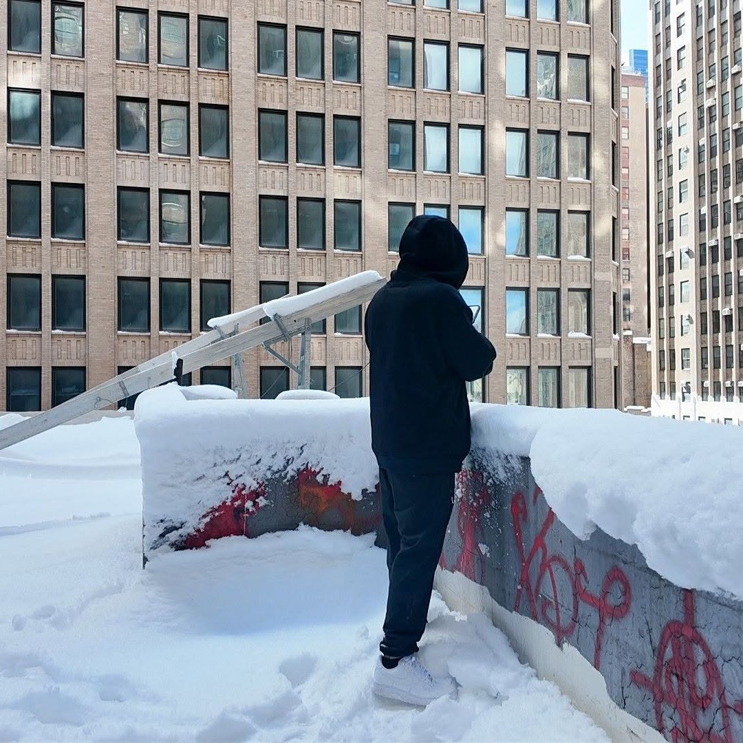 Standing on a snow-covered Manhattan rooftop beside a ladder, brick towers rising on every side