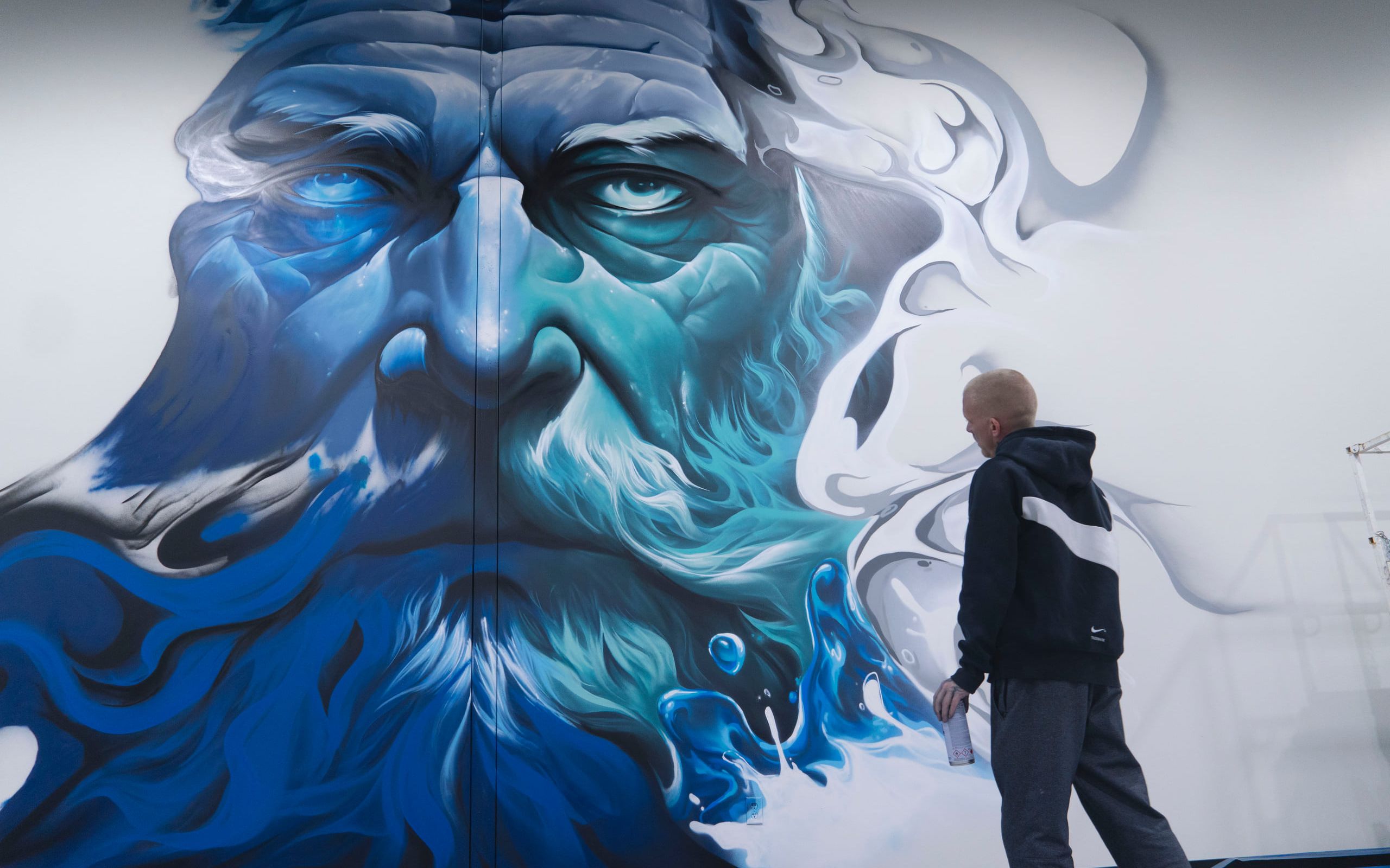 Monumental Zeus face mural in blue-toned grayscale, the artist standing before the nearly finished wall