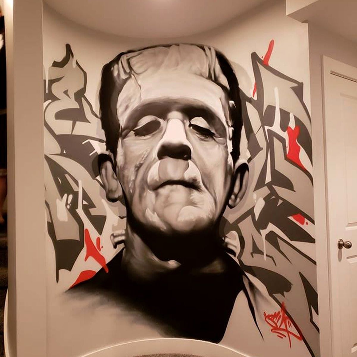 Black and white Frankenstein's monster portrait mural with red graffiti lettering in a hallway