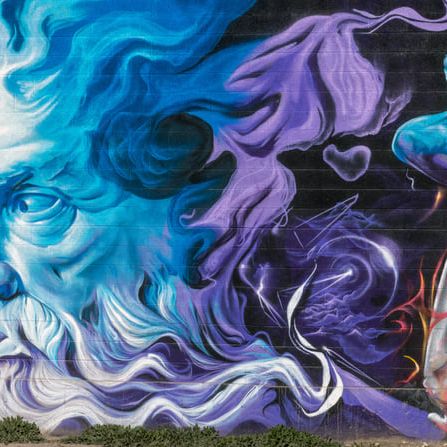 A long cinder-block wall painted end to end: bearded blue god, purple smoke, a falling figure in fire, and a giant eye