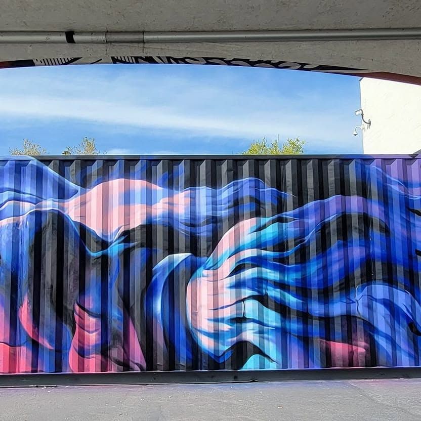 A bearded Greek god painted lying on his side along the ribbed steel of a shipping container, hair and beard streaming in blue and pink