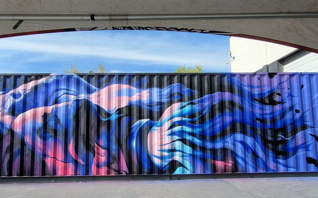 A bearded Greek god painted lying on his side along the ribbed steel of a shipping container, hair and beard streaming in blue and pink
