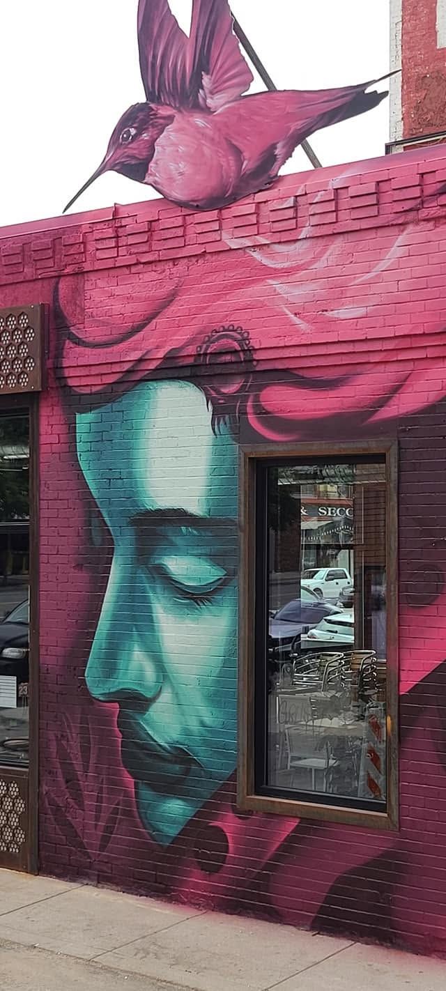 Teal portrait in profile with eyes closed, a magenta hummingbird above, painted on brick beside a window
