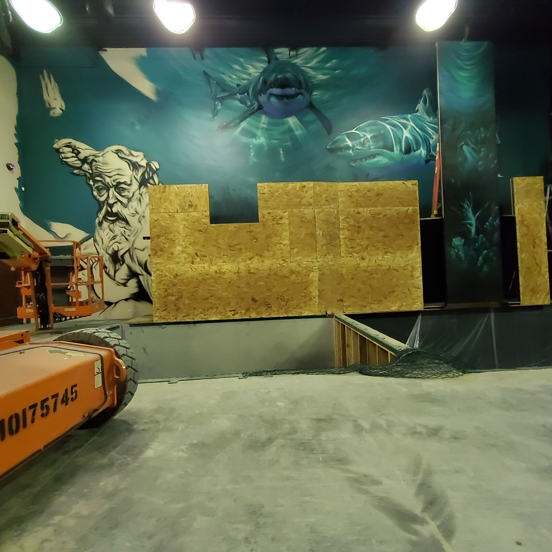 Half-painted 40ft wall of sharks and a marble god, with a boom lift and stacked sheeting on the bare concrete floor