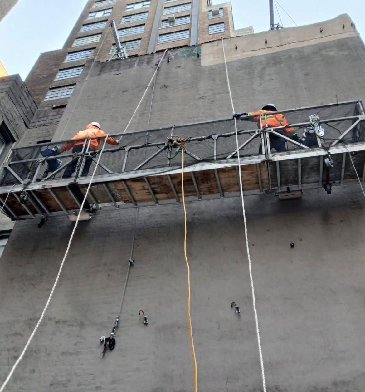 Two workers on a suspended scaffold rigged against a bare four-story side wall, seen from the street