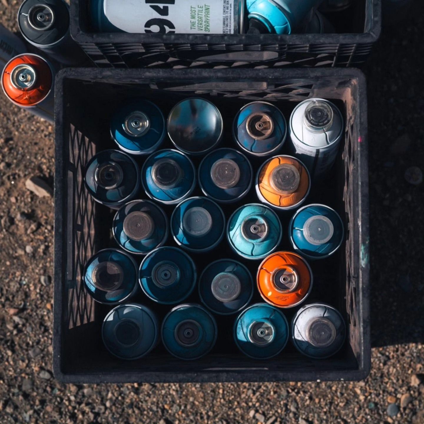Overhead view of a crate of spray-paint cans, caps up, on gravel beside a job site